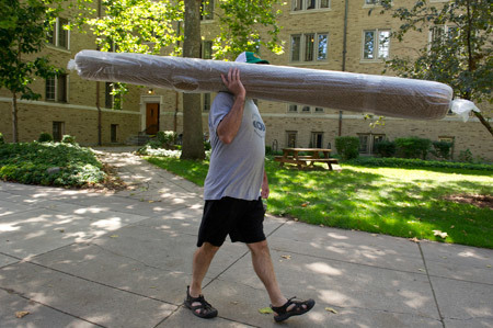 A father carries a rolled carpet across campus