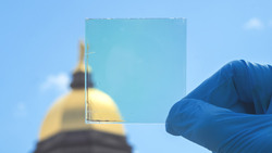 A latex gloved hand holds a small square of glass featuring a transparent coating in front of Notre Dame's golden dome.