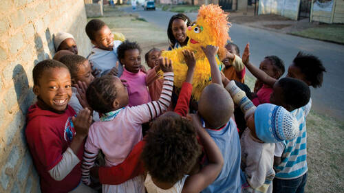 South African children play with Kami, a furry, yellow, five-year-old HIV-positive Muppet girl orphaned by AIDS.  ©2007 Sesame Workshop.  All rights reserved.  Photographed by Ryan Heffernan.