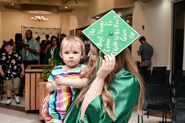 A graduate of the Goodwill Excel Center wearing a green cap and grown holds a little girl.