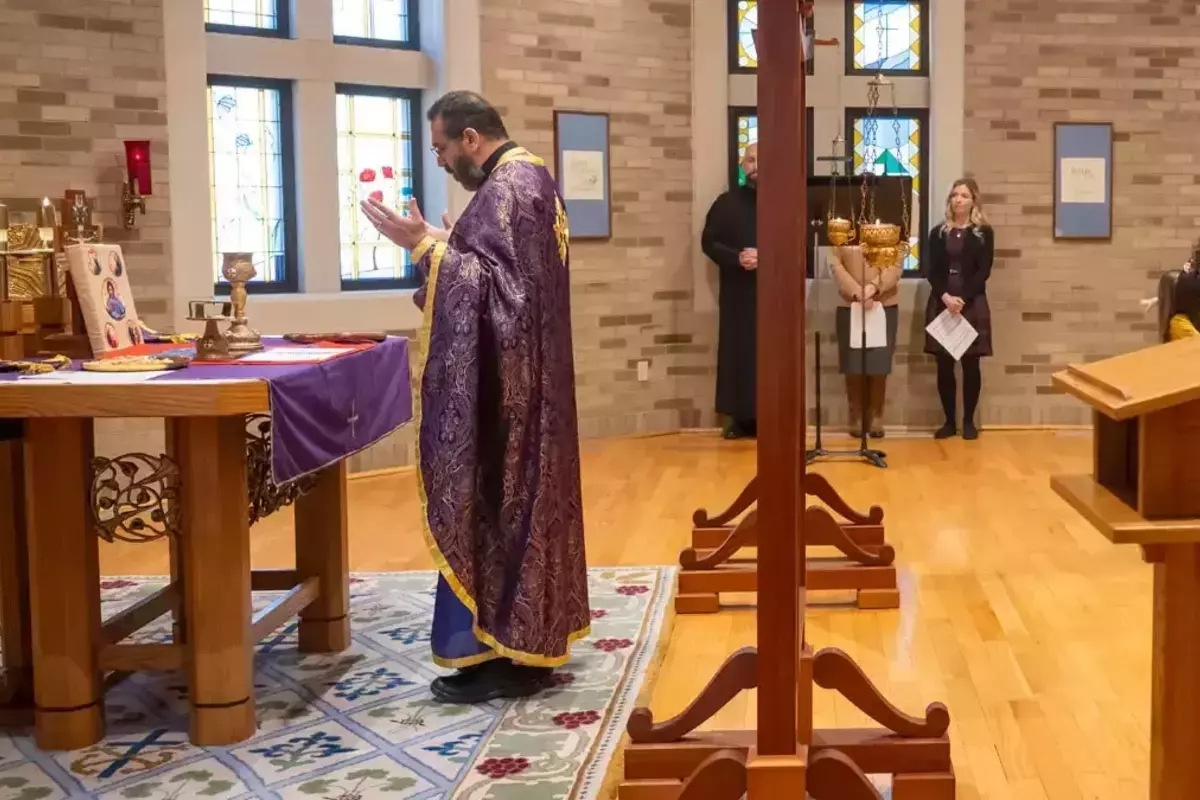 Fr. Khaled, wearing purple robes, stands in front of an altar with his arms up in prayer.