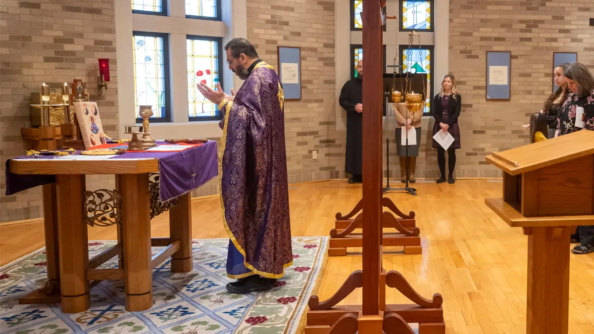Fr. Khaled, wearing purple robes, stands in front of an altar with his arms up in prayer.
