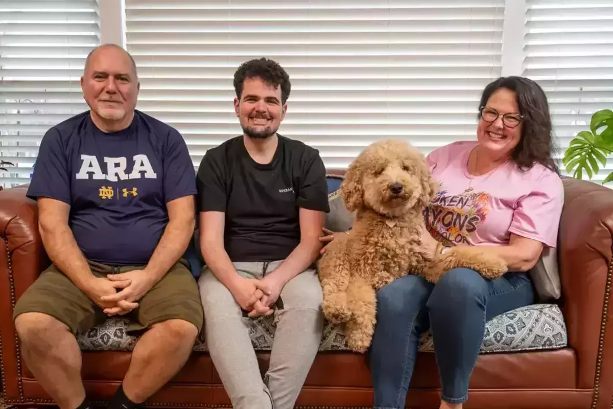 Harry, Alec and Gail Koujaian sit together on a brown leather couch with their goldendoodle dog.