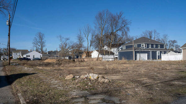 A view looking north across a vacant, dirt covered lot. It is late winter. Single-family homes can be seen in the background.