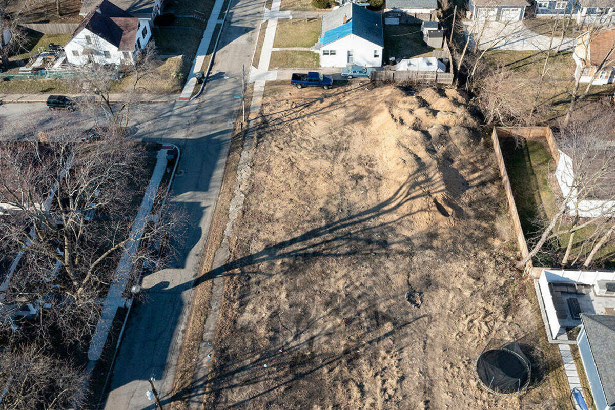 Aerial view of a vacant, dirt-covered lot surrounded by single-family homes. Barren trees cast long shadows across the lot. It is late winter.