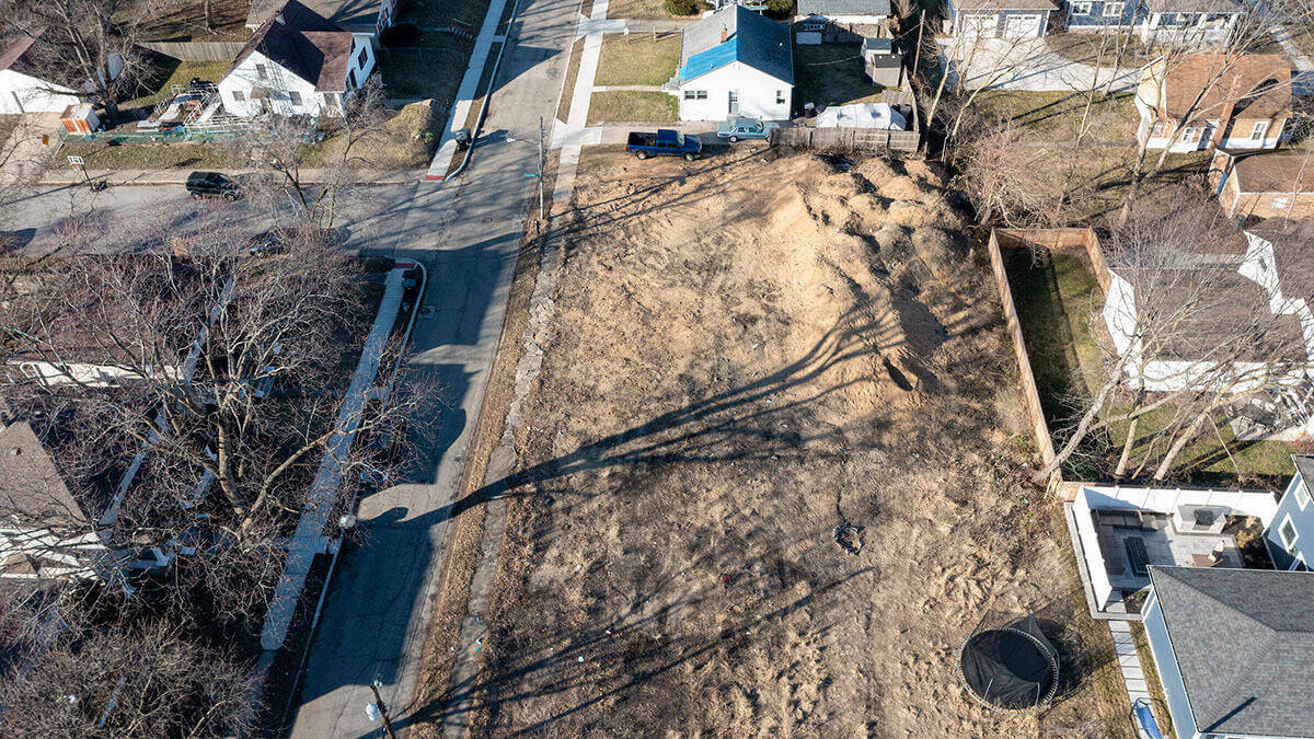 Aerial view of a vacant, dirt-covered lot surrounded by single-family homes. Barren trees cast long shadows across the lot. It is late winter.