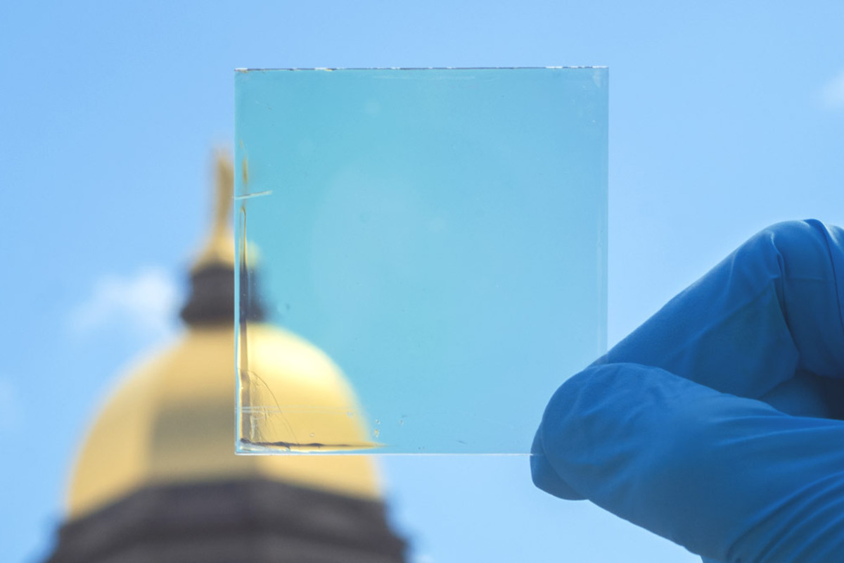 A latex gloved hand holds a small square of glass featuring a transparent coating in front of Notre Dame's golden dome.