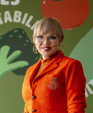 A woman with short hair ang glasses, hoop earrings and an orange blazer.