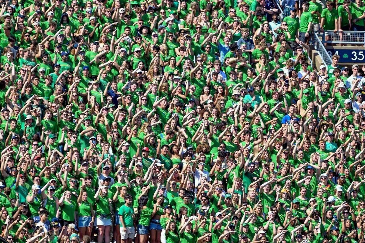 Hundreds of students in Notre Dame Stadium waving and mostly wearing green t-shirts on a sunny gameday.