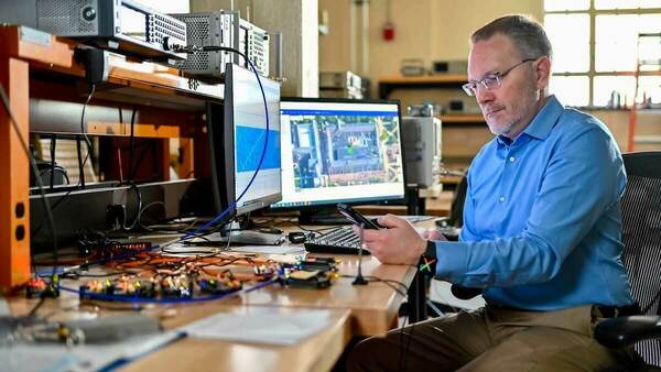 Nick Laneman, professor at Notre Dame and director of SpectrumX is pictured sitting at a desk in front of a computer and several electrical components. He is wearing glasses and a blue button up shirt.