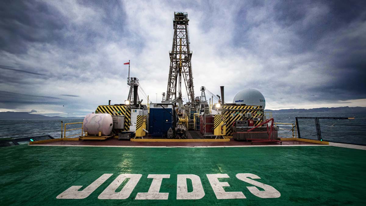 An image of the JOIDES Resolution, a 470-ft research vessel, while at sea. The word JOIDES is painted in white letters on a green deck. The sky above is cloudy.