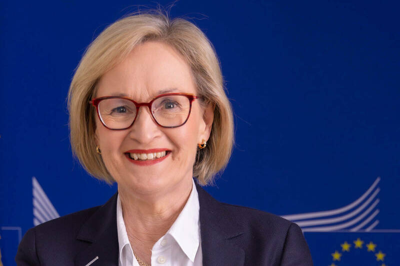 Mairead McGuinness is shown wearing a white shirt and dark blue blazer, with a short blond bob and red-rimmed eyeglasses.