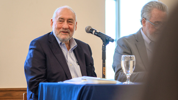 Joseph Stiglitz sits at a table with Ray Offenheiser beside him, and with a microphone in front of him for a lecture or discussion.