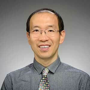 Xin Lu is pictured seated in front of a gray background. He has short dark hair, wears glasses and is wearing a gray shirt and dark tie.