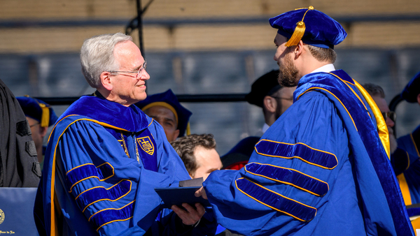 A man hands a diploma to another man during a commencement ceremony. The men wear matching blue robes. The man receiving the diploma also wears a blue cap with a gold tassel.