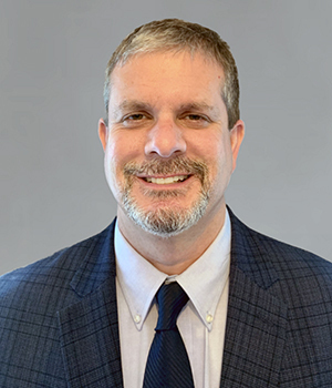 Professor Layman has salt-and-pepper colored hair, mustache and beard, and wears a dark blue blazer and tie over a white shirt.
