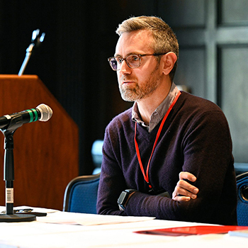 Professor Jaros sits at a table at a seminar with a microphone in front of him, presumably answering questions on a panel, with a podium in the background.
