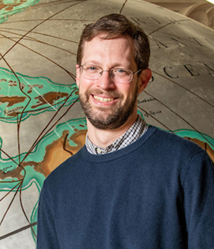 Professor Hungerman stands in front of a large image of a globe, wearing a blue sweater and glasses.