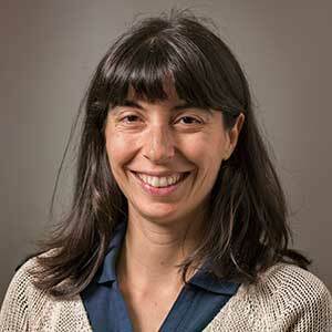 Paola Crippa smiles in front of a gray backdrop. She has long dark hair and bangs and is wearing a blue shirt under a beige sweater.