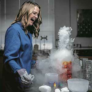 Kate Biberdorf is pictured smiling in a laboratory. A white cloud rises from a beaker. Kate is wearing a lab coat and protective glasses.