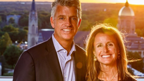 A well-dressed couple poses for a photo with the University of Notre Dame campus in the background.