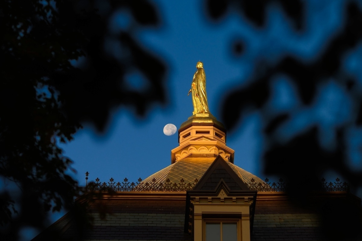 Golden dome illuminated at nighttime, against a dark blue sky with the moon to the left.