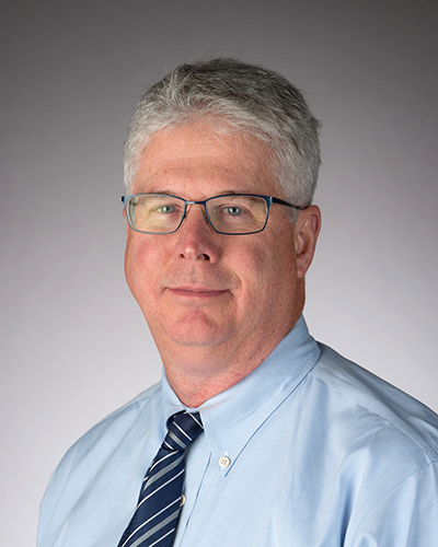 Professor Bill Evans has gray hair, thin-rimmed glasses, and is wearing a light blue shirt with dark blue tie.