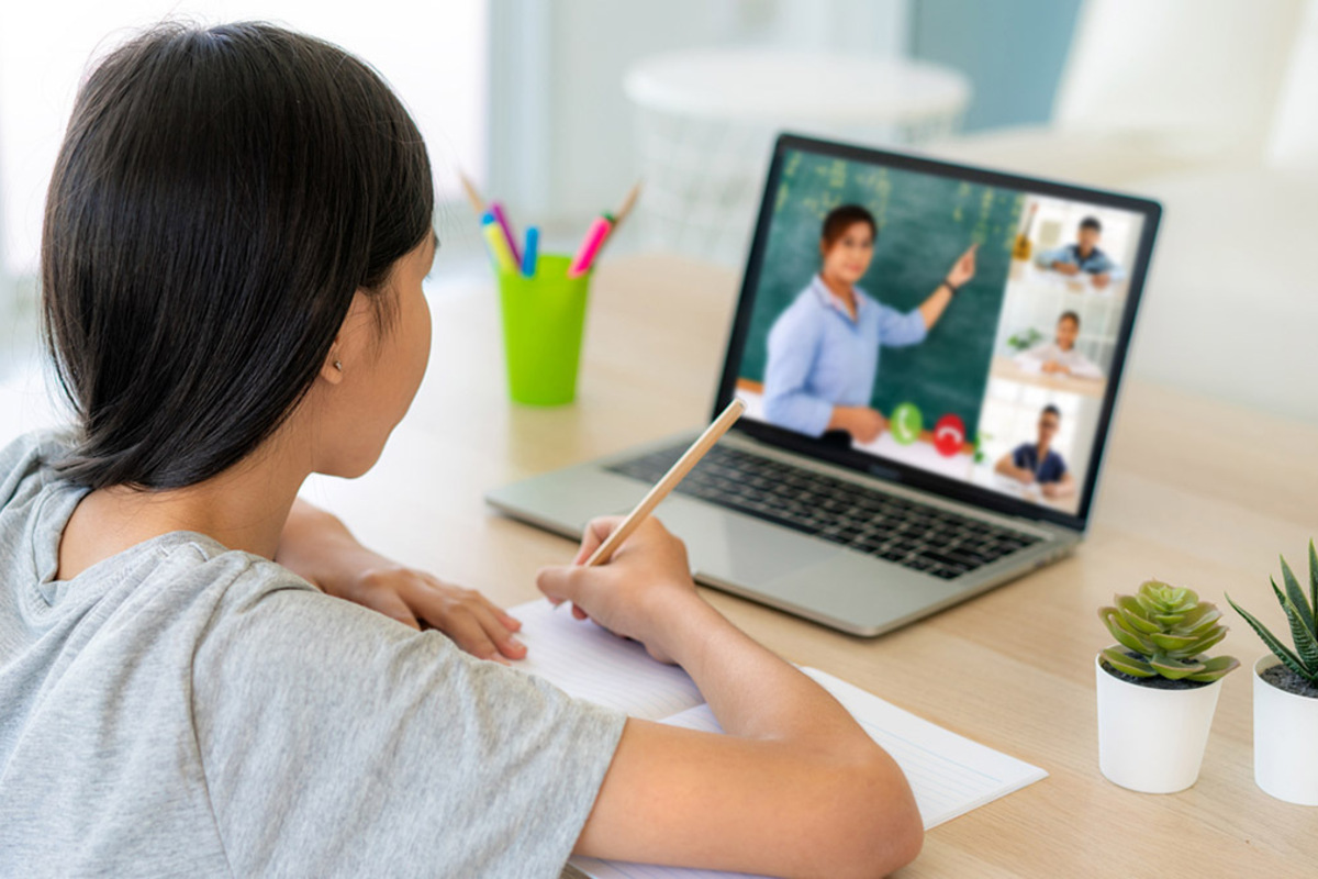 Young middle school student with long black hair sits at her desk in front of a laptop where you can see on the screen that a teacher is giving virtual instruction.