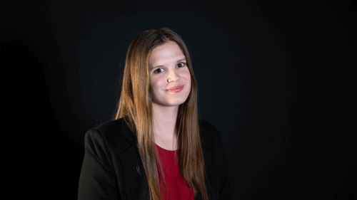 A headshot of a young woman with long brown hair against a black background. She wears a black blazer and a dark red shirt. She is smiling slightly and looking off to the right side of the frame.