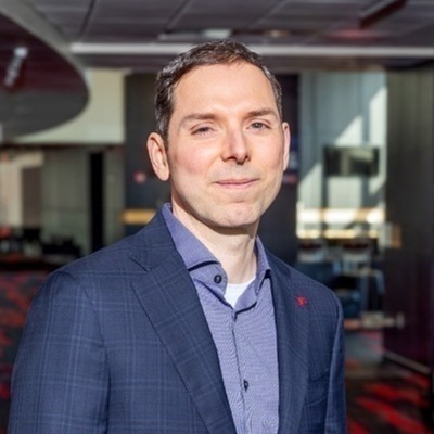 A man with short brown hair and a slight smile wears a blue plaid suit jacket over a purple button-down shirt with a red pin on his lapel, standing in a modern indoor setting.