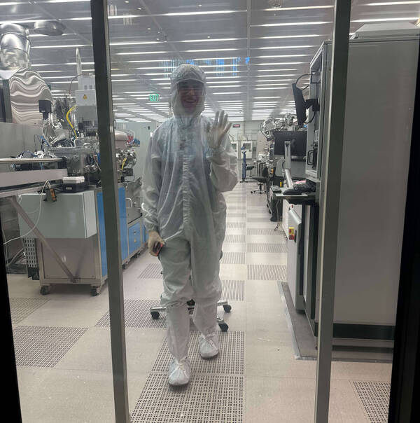 Smiling person in a white cleanroom suit, face shield, and gloves waves from a lab with machinery.