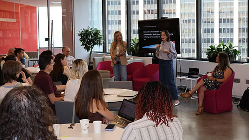Woman presents to an audience in a modern room with large windows.